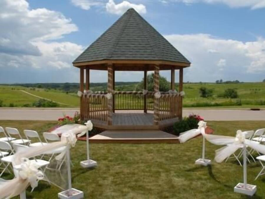 A gazebo with a sloped roof, surrounded by white chairs and tables, set against a rural landscape.