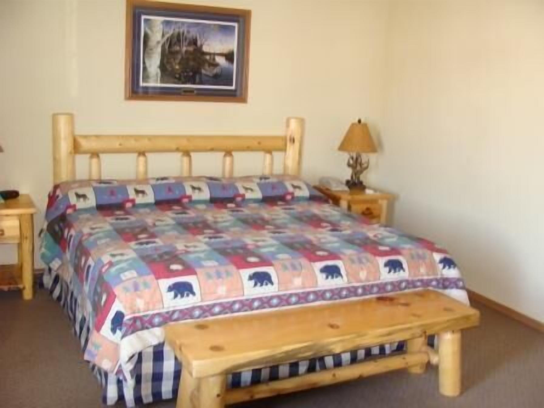 A bedroom with a wooden bed frame, a quilt with animal prints, a bedside table with a lamp, and a framed picture on the wall.
