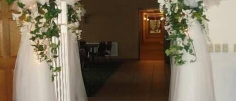 Un arco interior con cortinas blancas y follaje, que conduce a una habitación con suelo de madera y una zona de comedor al fondo.