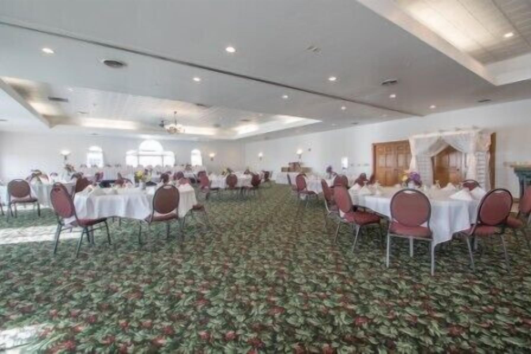 A large banquet hall with round tables and chairs, carpeted floor, and a high ceiling with recessed lighting.