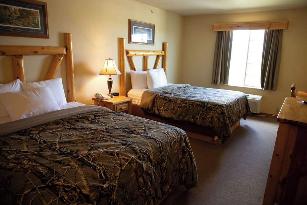 A hotel room with two beds, wooden headboards, a nightstand with a lamp, a framed picture on the wall, and a window with curtains.