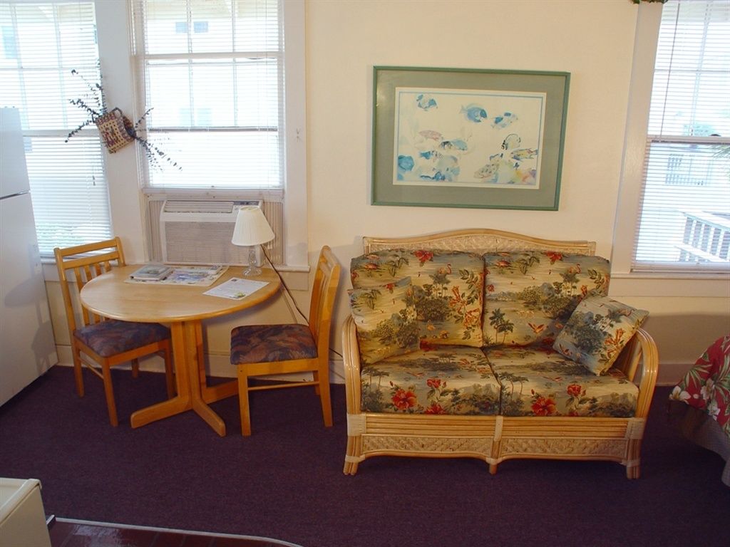 A room with a floral sofa, a round wooden table with chairs, a framed artwork on the wall, and a window with blinds.