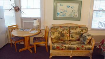 A room with a floral sofa, a round wooden table with chairs, a framed artwork on the wall, and a window with blinds.