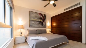 A bedroom with a bed, two bedside lamps, a ceiling fan, a large abstract painting, and a wooden wardrobe.