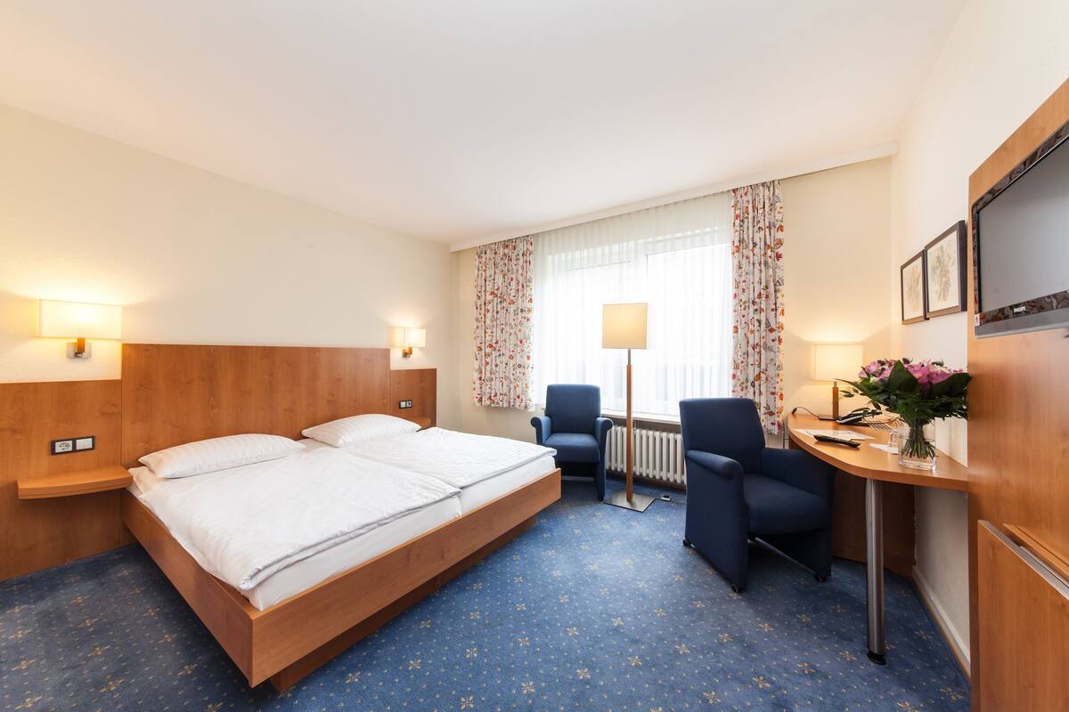 A hotel room with a large bed, two bedside tables, a desk with a chair, a radiator, and a window with curtains.