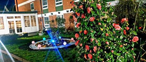 Un jardín con una fuente y un rosal frente a un edificio con múltiples ventanas.