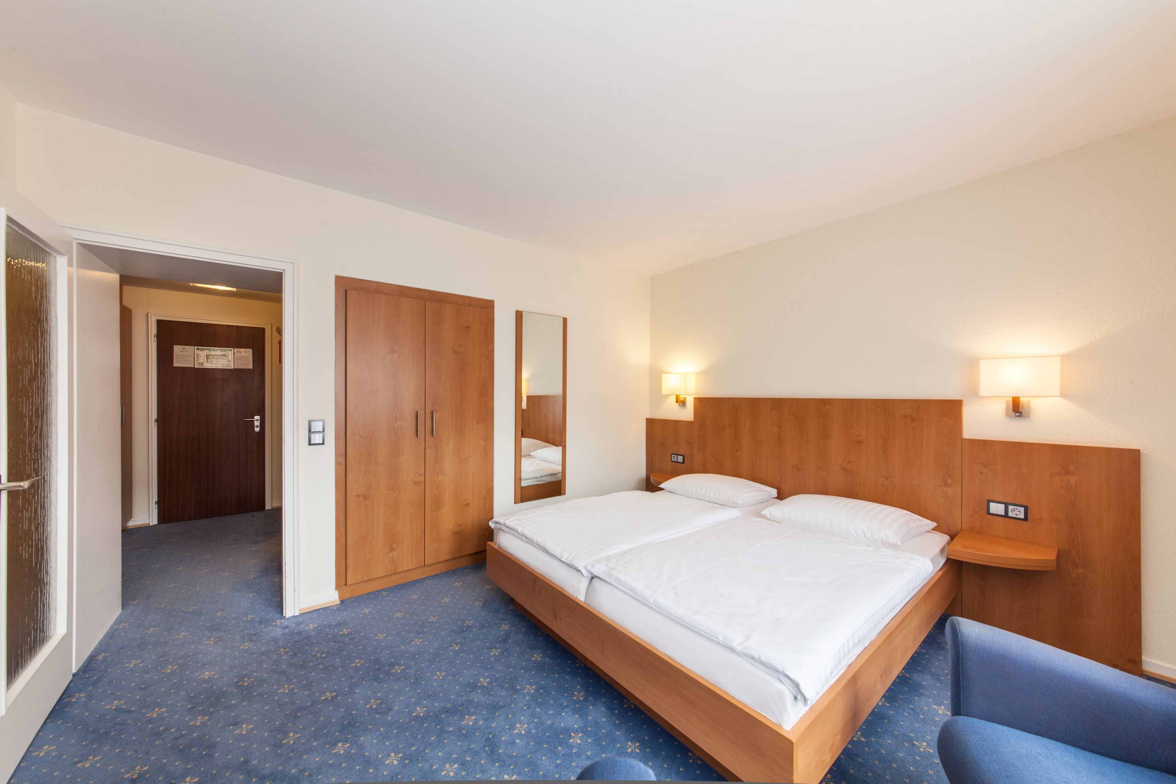 A hotel room with a large bed, wooden headboard, two bedside tables with lamps, a wardrobe, and a mirror.