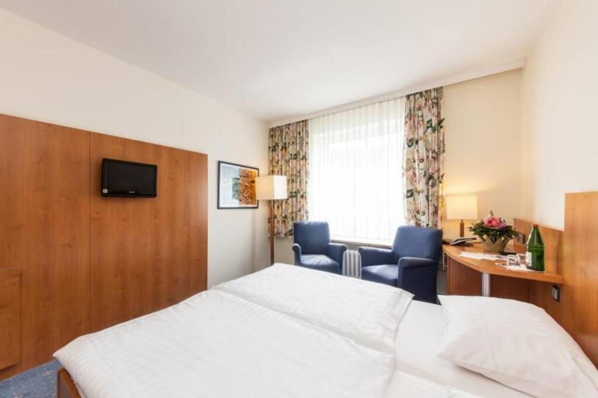 A hotel room with a bed, two chairs, a desk with a lamp, a television, and a window with curtains.