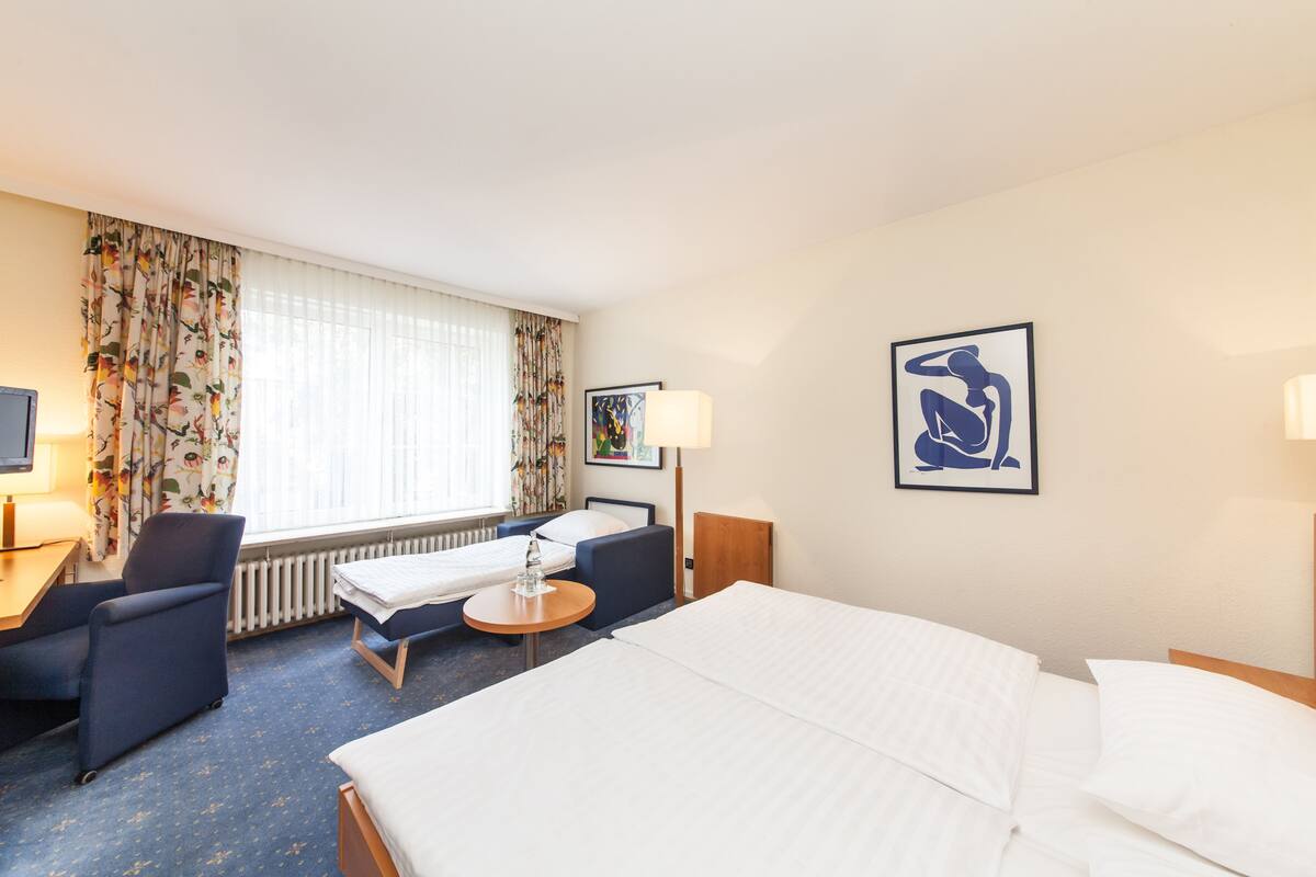 A hotel room with a large bed, a desk with a chair, a small table, and a window with curtains.