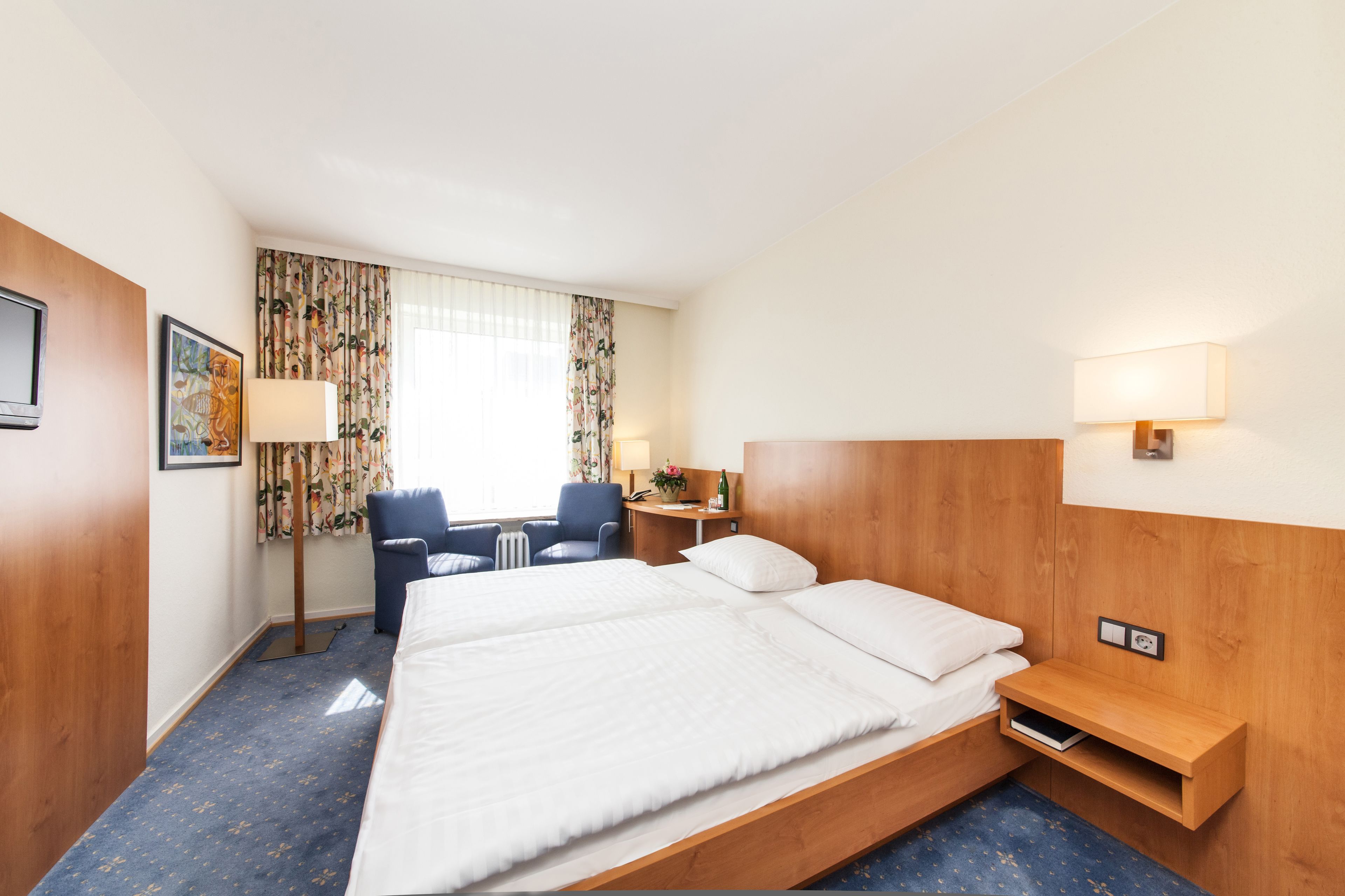 A hotel room with a large bed, a desk, a chair, a lamp, and a window with curtains.