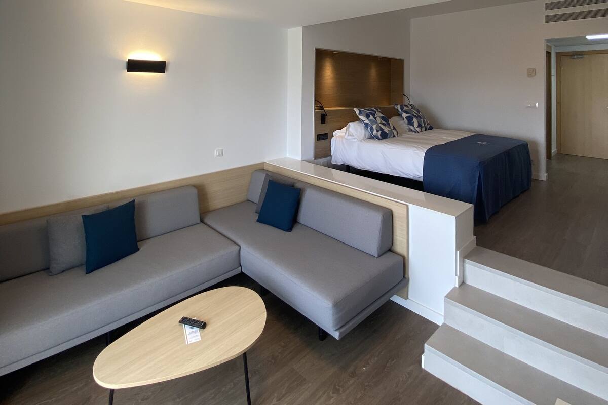 Ein modernes Hotelzimmer mit einem grauen Ecksofa, einem kleinen runden Tisch, einem Bett mit blauer Bettdecke und einer Treppe, die zu einer weiteren Ebene führt.