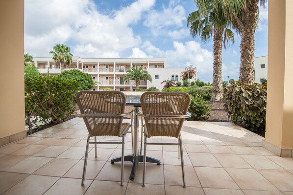 A patio with two wicker chairs and a table, overlooking a building with balconies and palm trees.