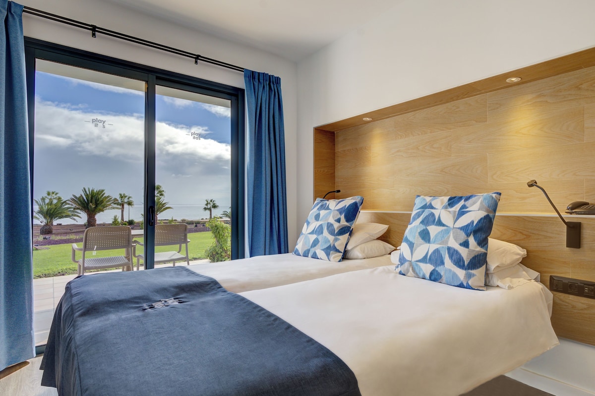 Habitación de hotel con dos camas, un ventanal con cortinas, cabecera de madera y vistas al exterior.