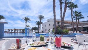 Un espace repas aménagé au bord de la piscine, avec une table dressée pour deux personnes, proposant une variété de plats et de boissons, et offrant une vue sur une piscine et des palmiers.