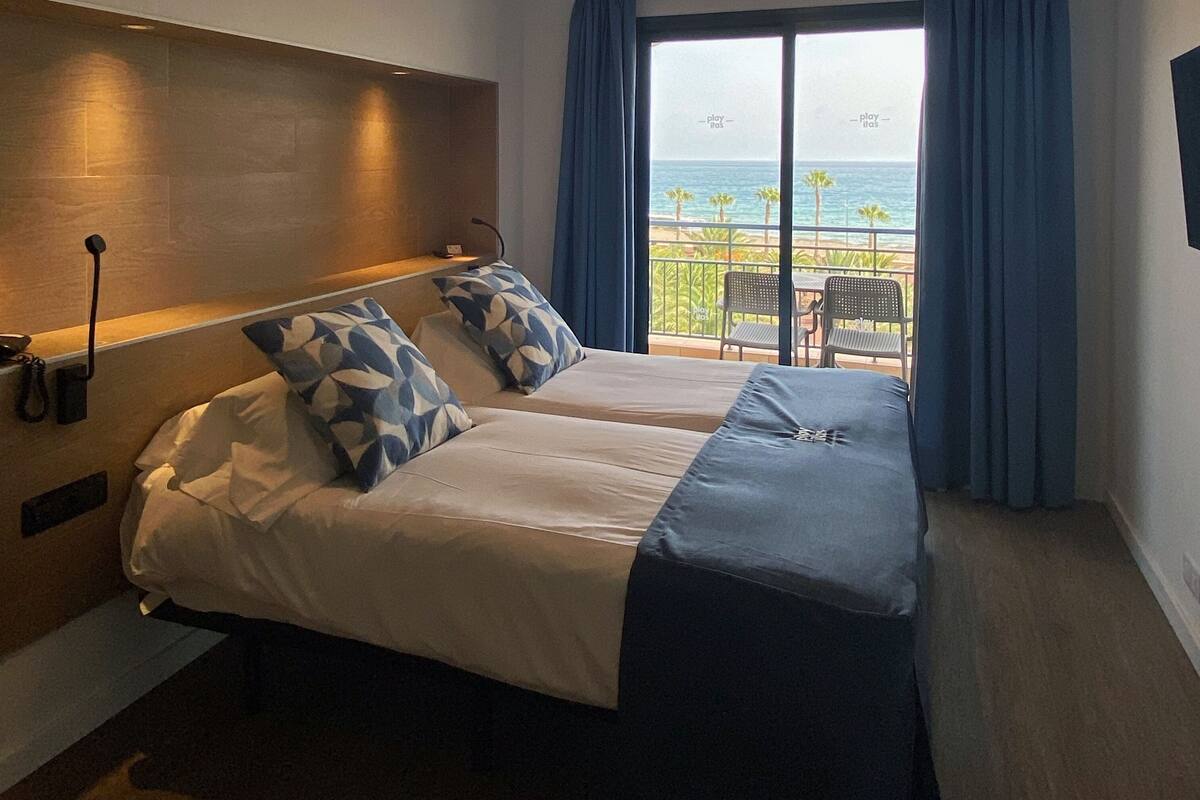 A hotel room with two beds, a balcony with a view of the sea, and a small table with chairs.