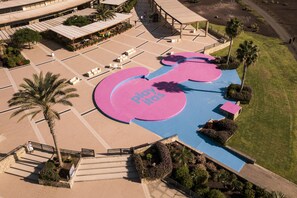 Luchtbeeld van een hotel met een centrale binnenplaats, palmbomen en een blauwe en roze speelruimte.