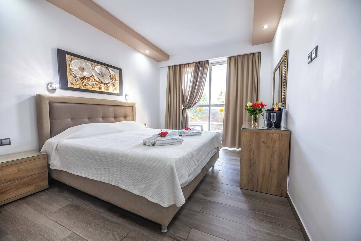 A modern bedroom with a large bed, a wooden nightstand, a bedside table, a mirror, and a window with curtains.