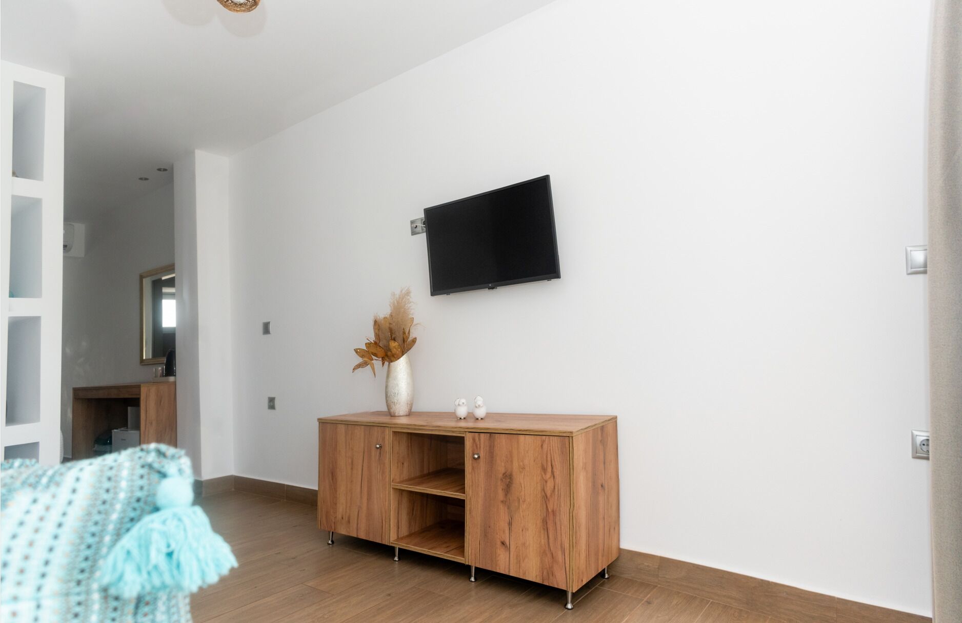 Una habitación moderna con un mueble de madera, un televisor de pantalla plana montado en la pared y un florero con flores secas sobre el mueble.