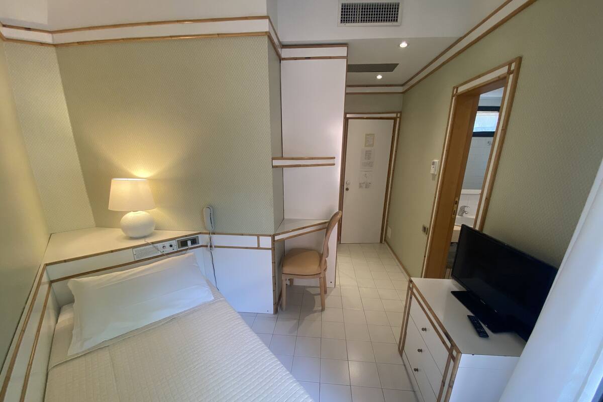 A hotel room with a bed, bedside table with a lamp, a TV, a desk with a chair, and a wardrobe.
