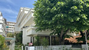 A modern hotel with a white facade, a large tree in front, and a white picket fence.