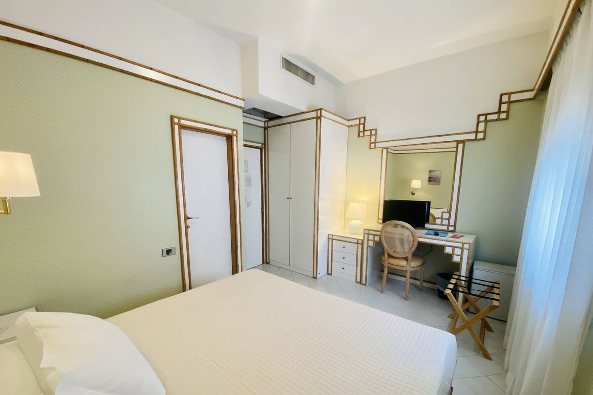 A hotel room with a bed, a desk with a chair, a computer, and a mirror.