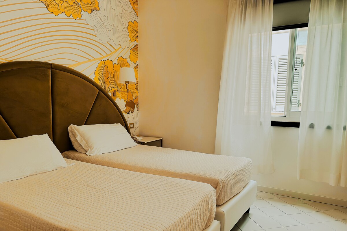 A hotel room with two beds, a large headboard, a nightstand, and a window with curtains.