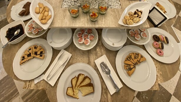 A dining table set with various food items, including pastries, fruit, and condiments, surrounded by chairs and a decorative plant centerpiece.