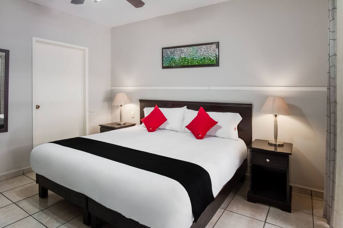 A neatly made bed with white and black bedding, two red pillows, two bedside lamps, and a framed picture on the wall.