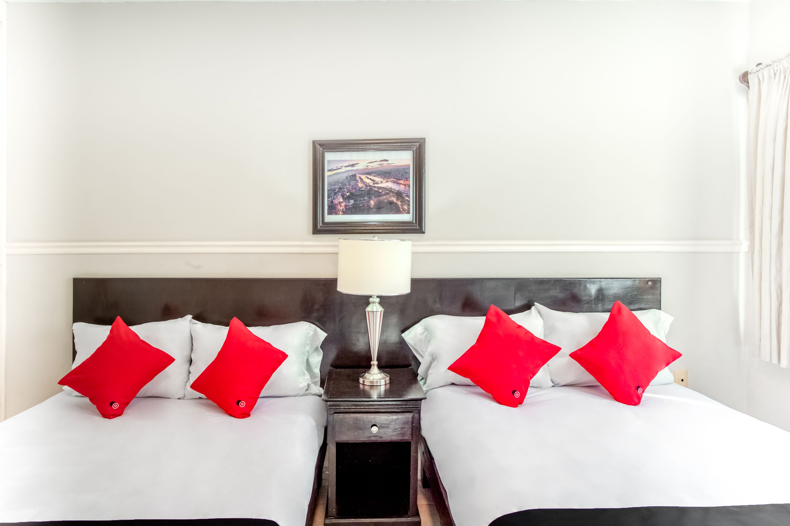 Habitación de hotel con dos camas, cada una con ropa blanca y almohadas rojas, una mesita de noche con lámpara y un cuadro en la pared.
