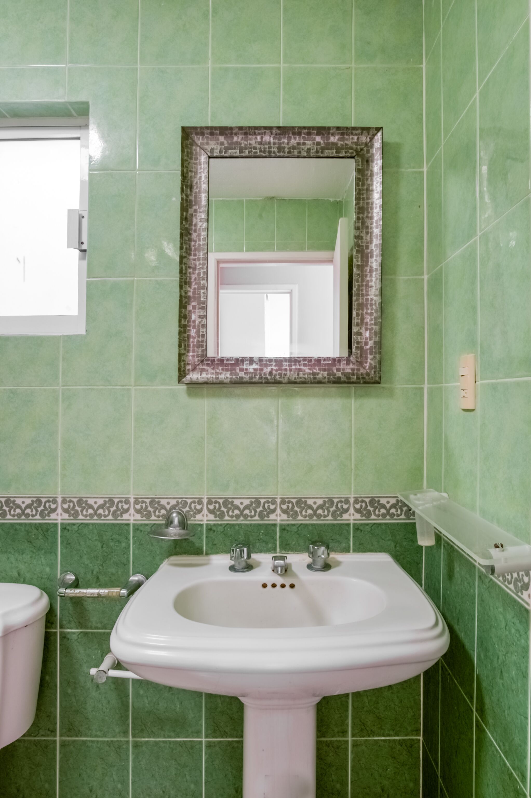 A bathroom with green tiles, a white sink, and a mirror with a decorative frame.
