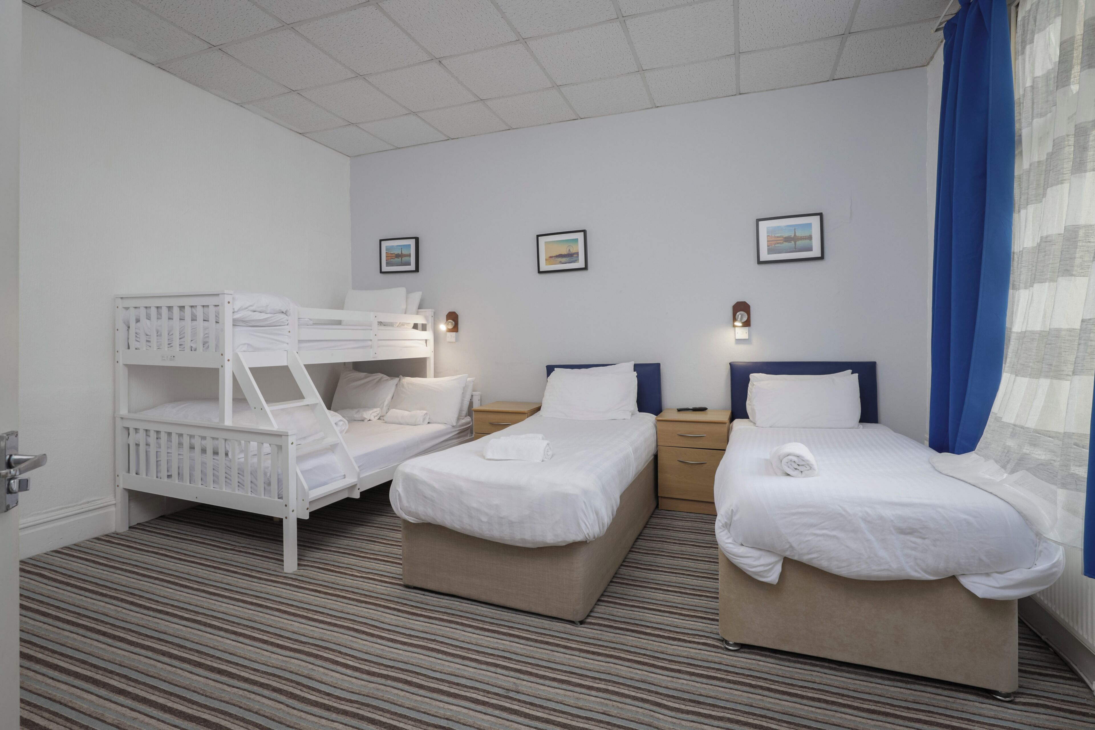 A hotel room with two beds, a bunk bed, a wooden nightstand, a wall-mounted lamp, and a window with blue curtains.