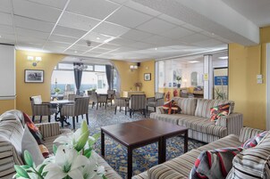 Un hall de hotel con sofás a rayas, una mesa de centro, un comedor con sillas y vistas a través de un ventanal.