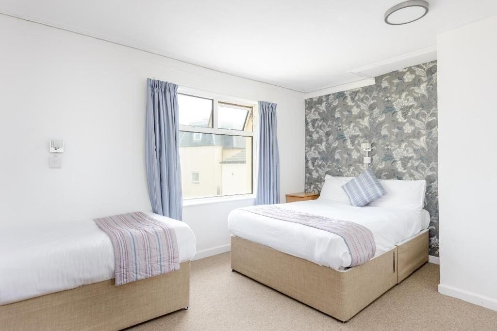 A bedroom with two beds, a window with curtains, a nightstand, and a wall with a floral pattern.