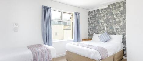Un dormitorio con dos camas, una ventana con cortinas, una mesita de noche y una pared con estampado floral.