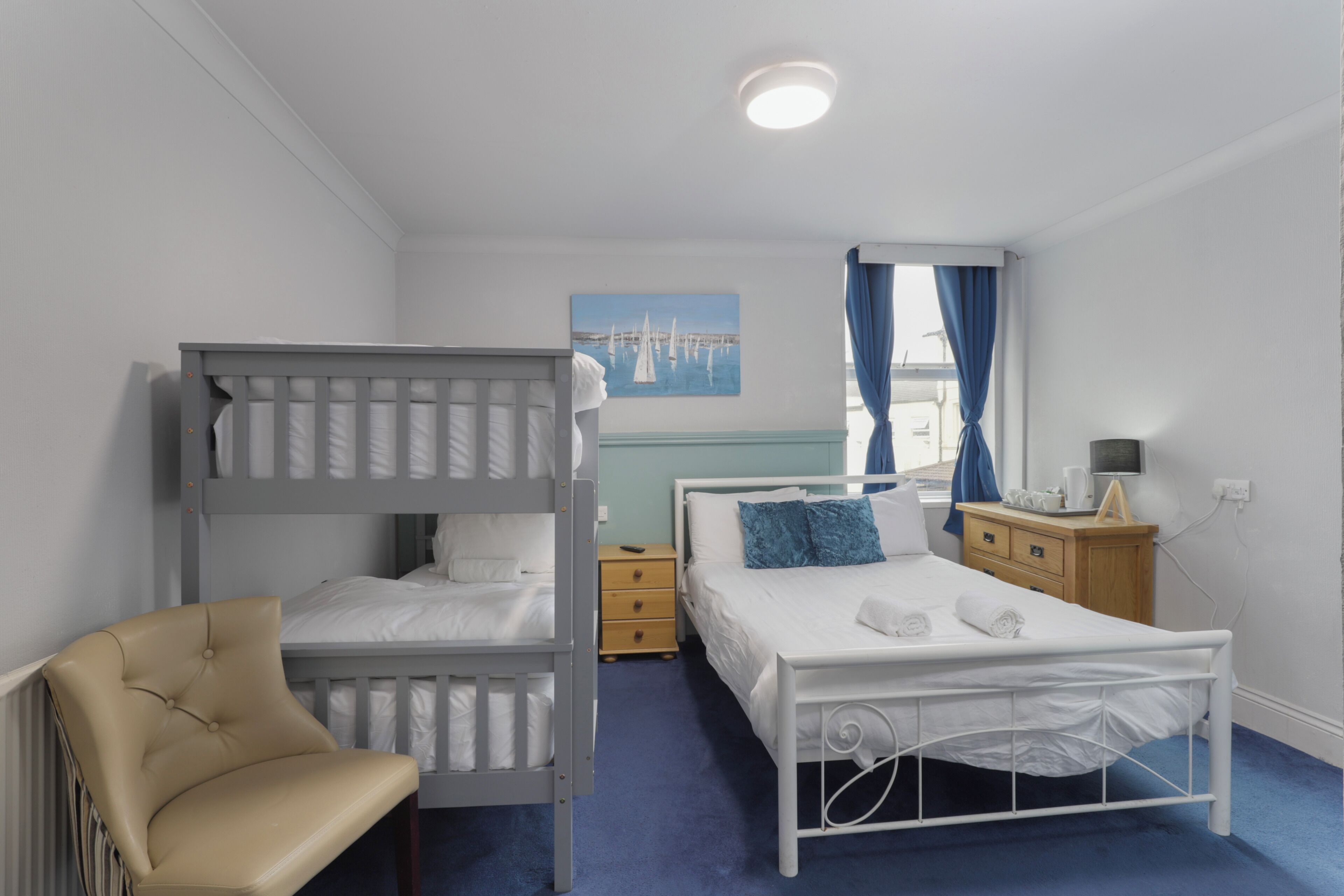 Une chambre avec des lits superposés, un lit simple, une table de chevet, une chaise et un tableau accroché au mur.