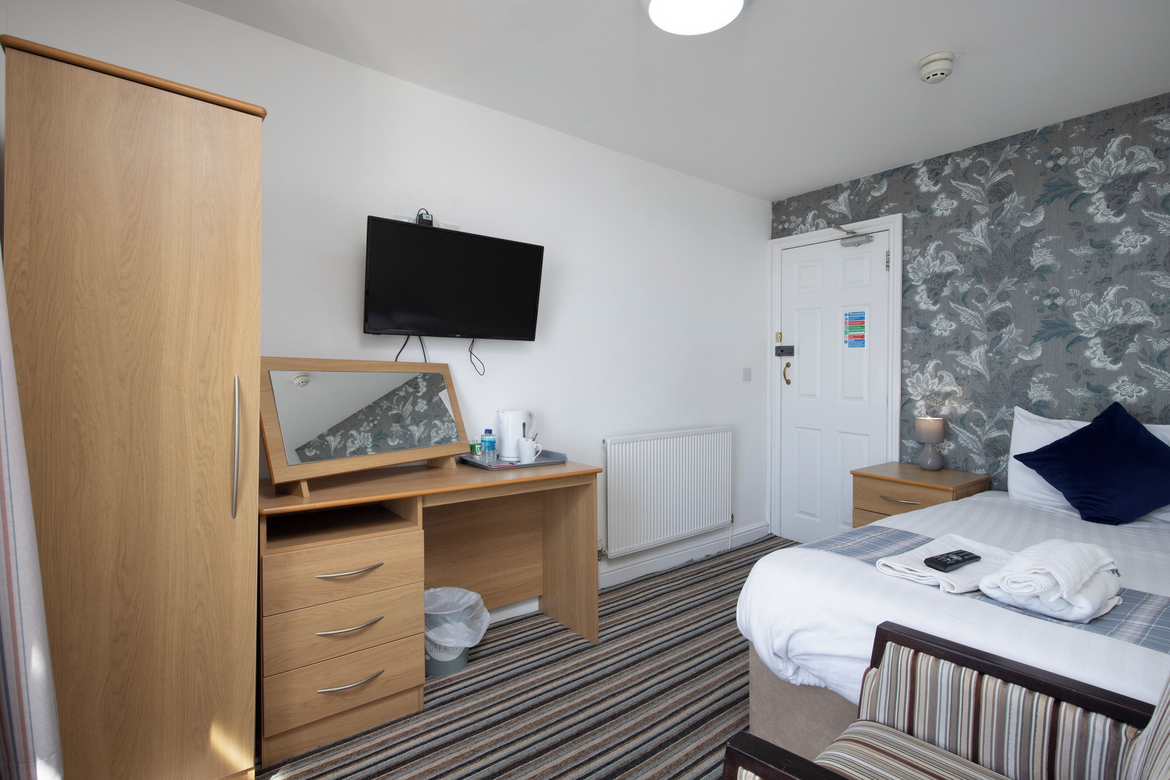 A hotel room with a wooden wardrobe, a desk with a mirror, a flat-screen TV, a bed with a striped pillow, and a radiator.
