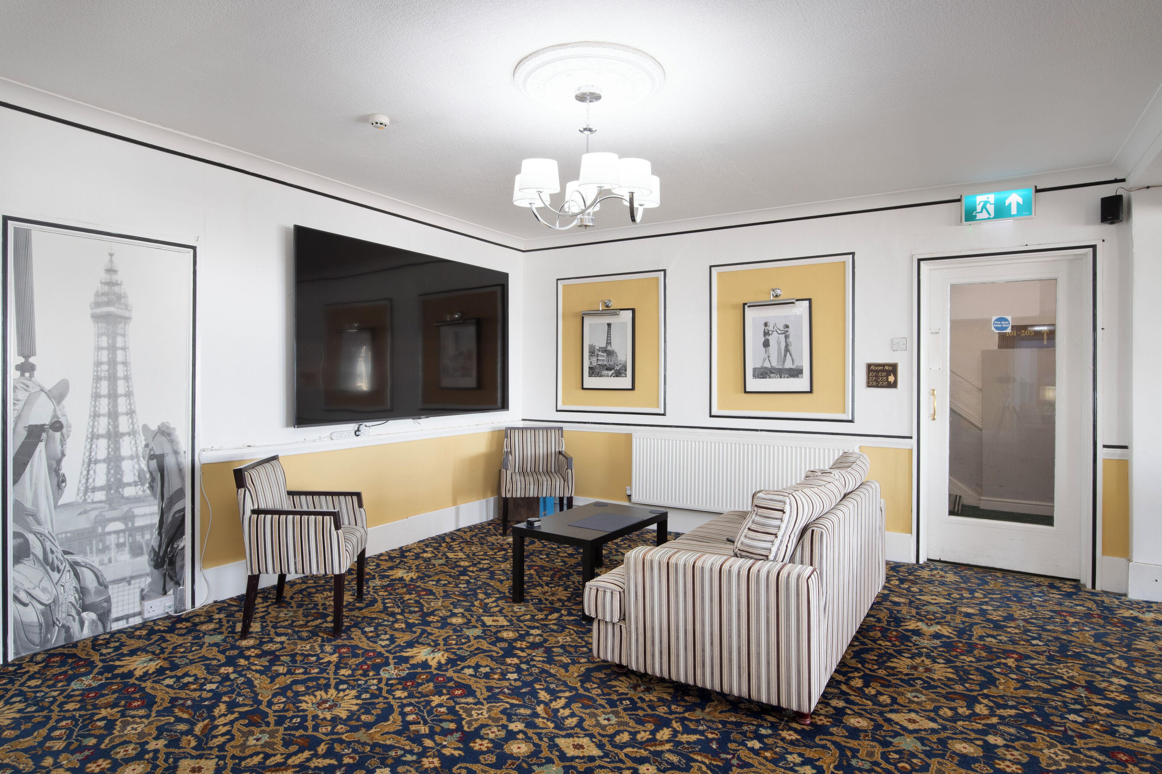 Eine Hotelhalle mit einem gemusterten Teppich, einer Couch, zwei Sesseln, einem Fernseher, gerahmten Bildern an den Wänden und einer Kronleuchter.