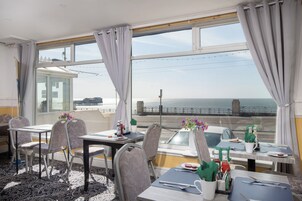 Comedor con mesas y sillas, vista al mar y una ventana con cortinas.