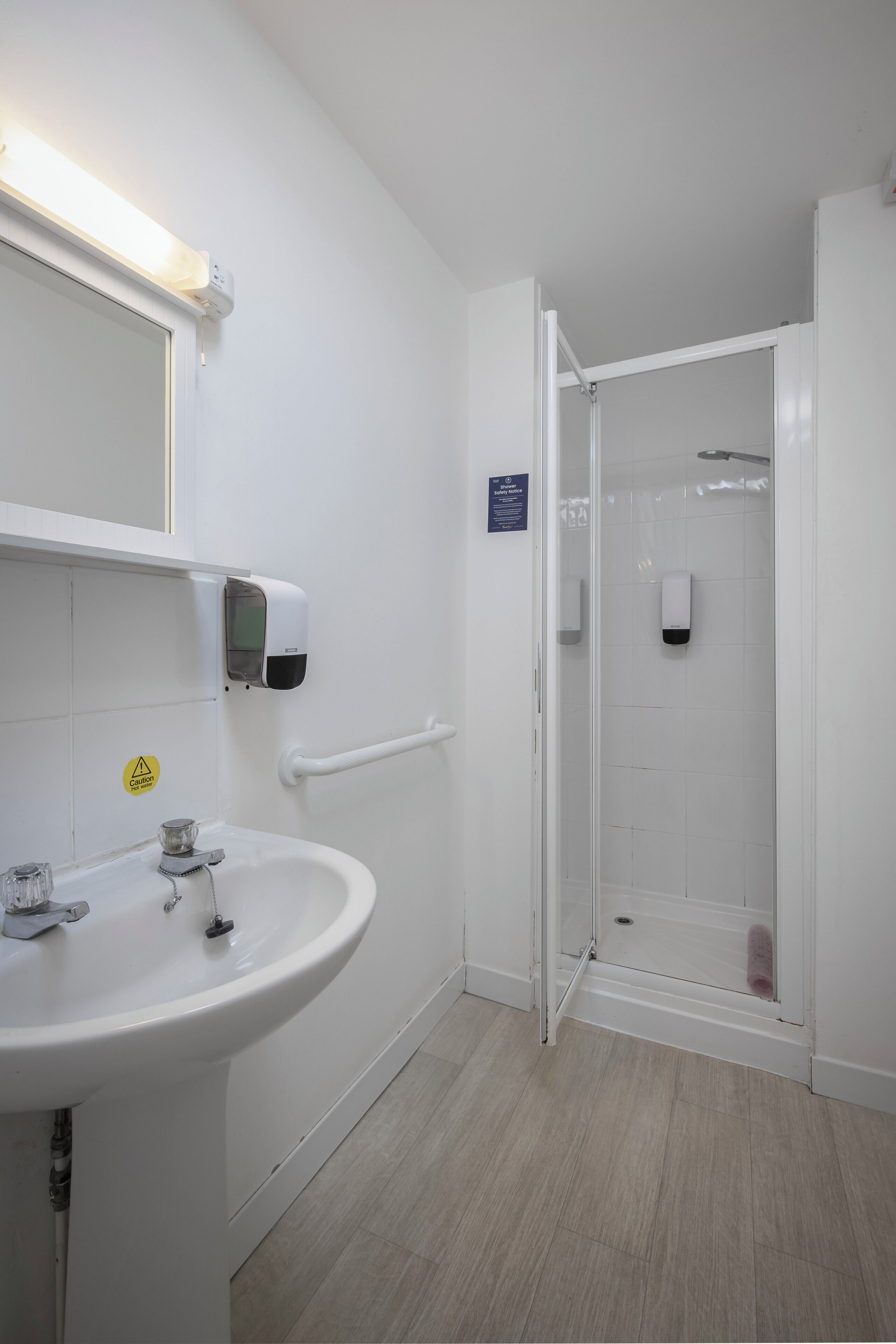 A bathroom with a white sink, a hand sanitizer dispenser, a glass shower enclosure, and a handrail.