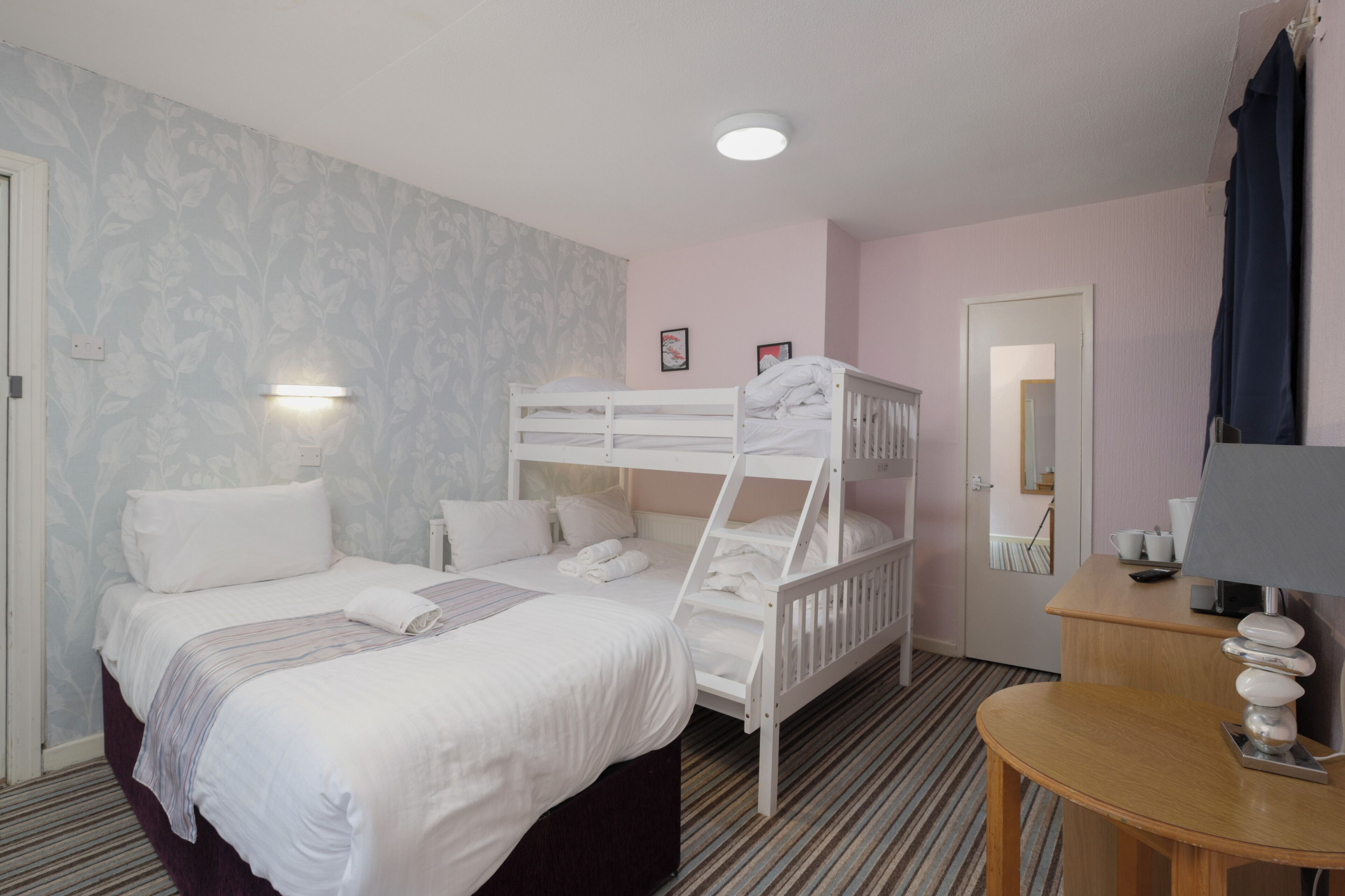 A hotel room with a bunk bed, a single bed, a bedside table with a lamp, a mirror, and a small desk with a cup and remote control.