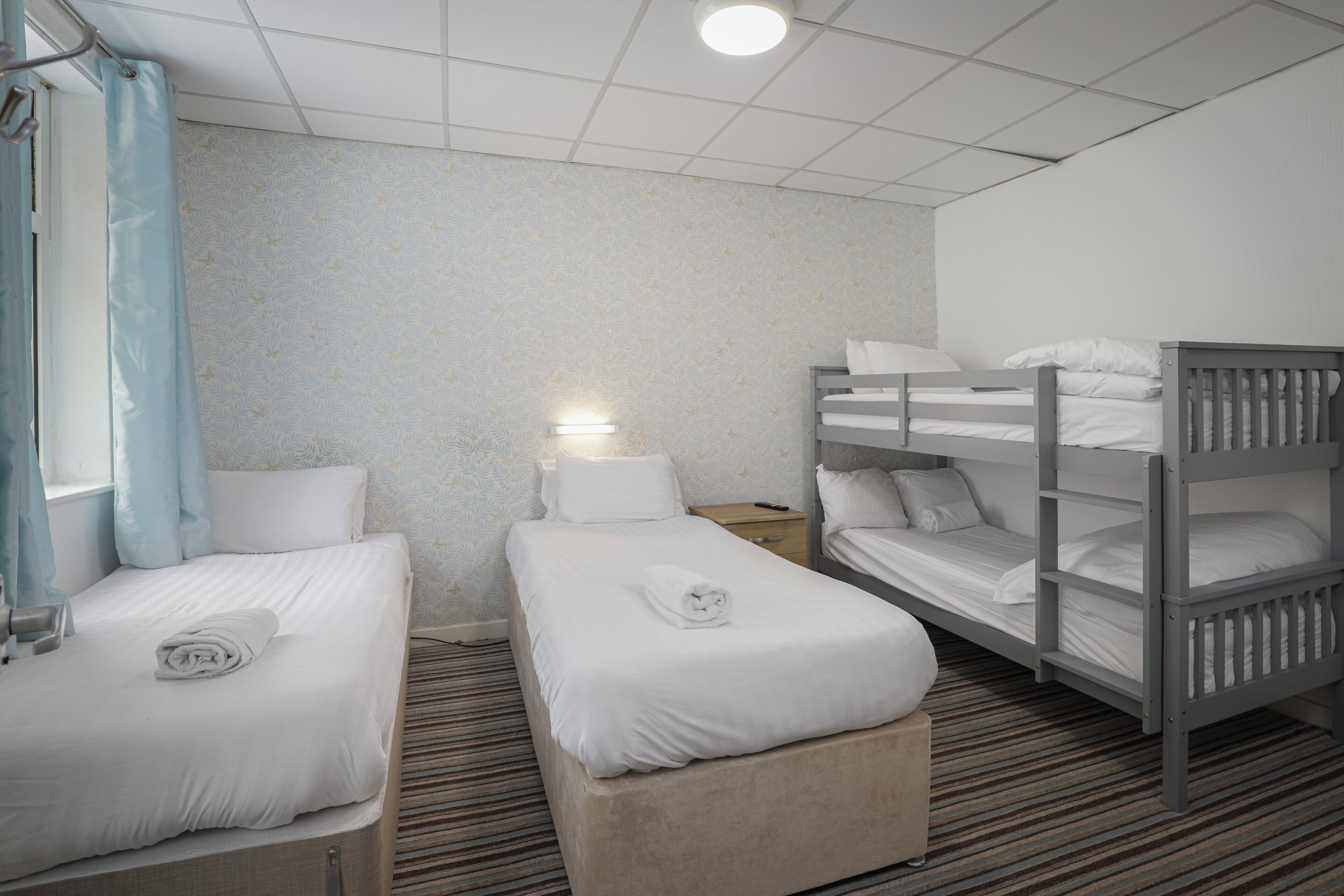 A hotel room with two single beds, a bunk bed, a bedside table with a lamp, and a striped carpet.