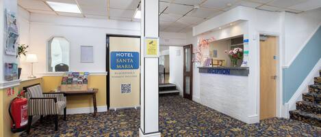 A hotel reception area with a reception desk, a fire extinguisher, a coat rack, and a wall-mounted sign for the hotel.