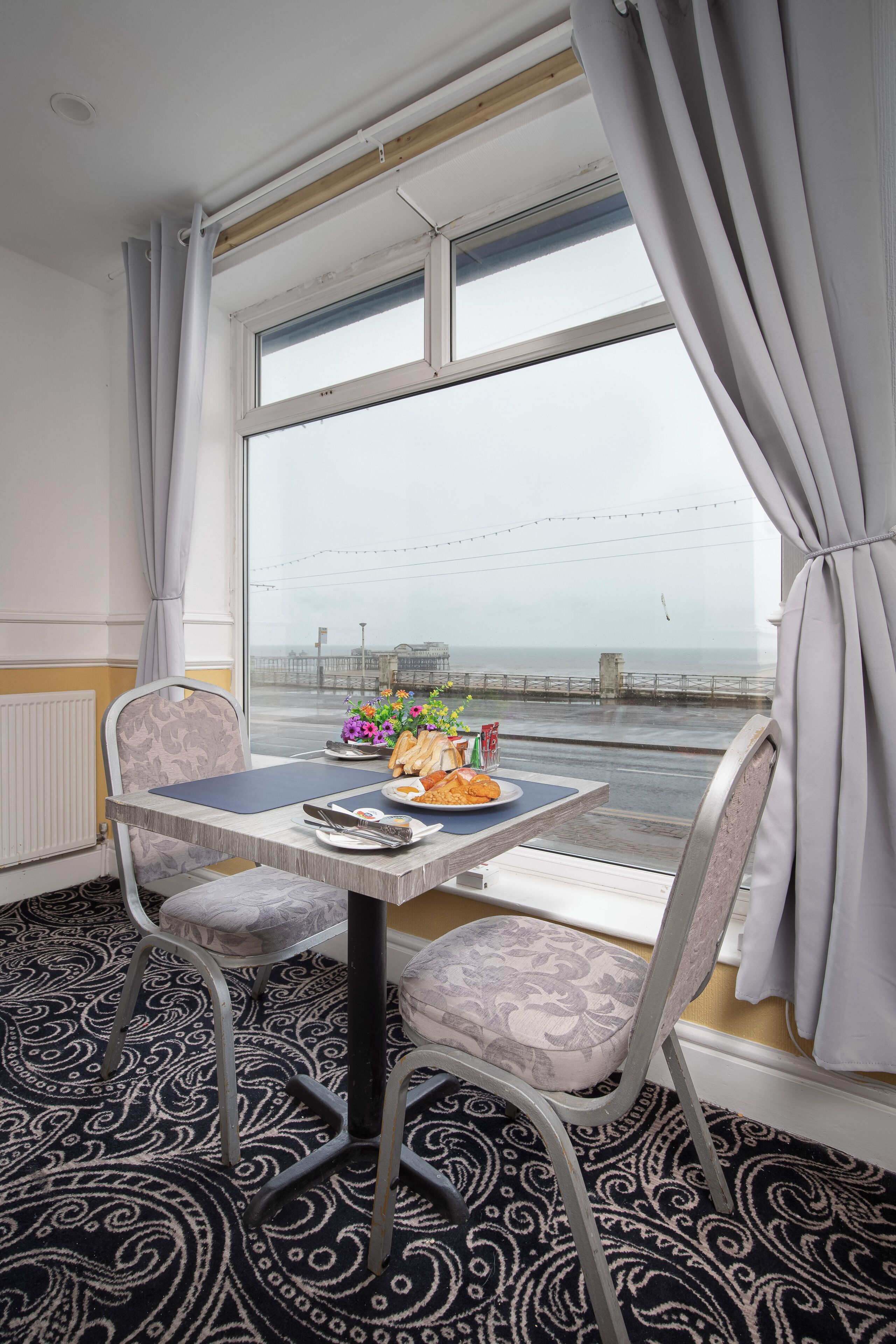 A room with a table set for breakfast, a chair, a window with curtains, and a patterned carpet.