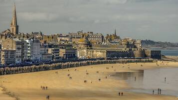 A coastal cityscape with historic buildings, a prominent church tower, and a sandy beach with people.