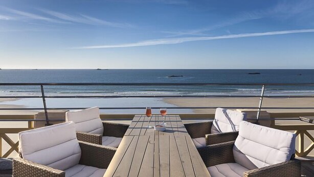 Uma mesa de madeira com duas cadeiras e dois copos de bebida numa varanda com vista para a praia.