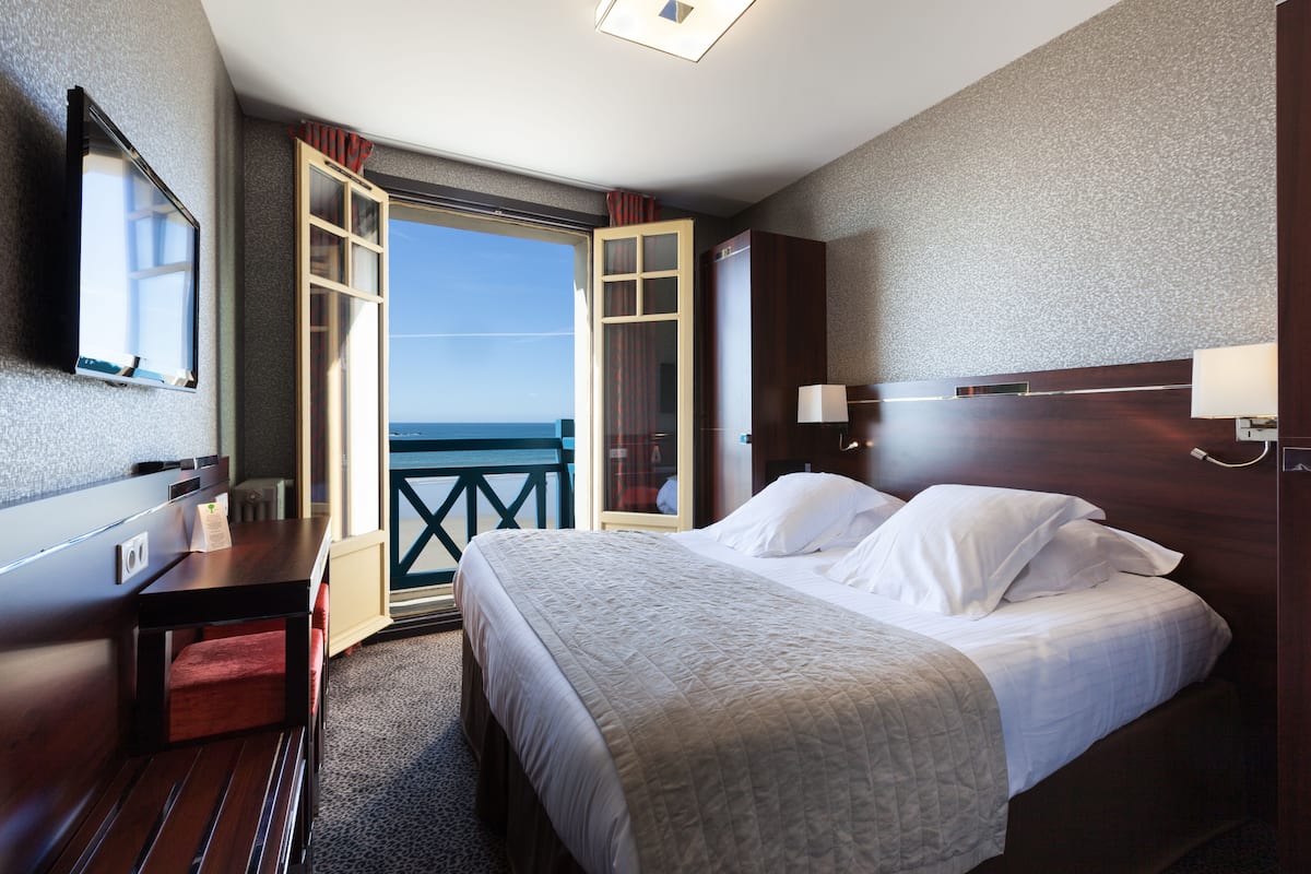 A hotel room with a bed, a desk, a chair, a TV, and a balcony with a view of the ocean.