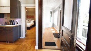 A modern kitchen with grey cabinets and a wooden floor, leading to a bedroom with a large window and a wooden door.