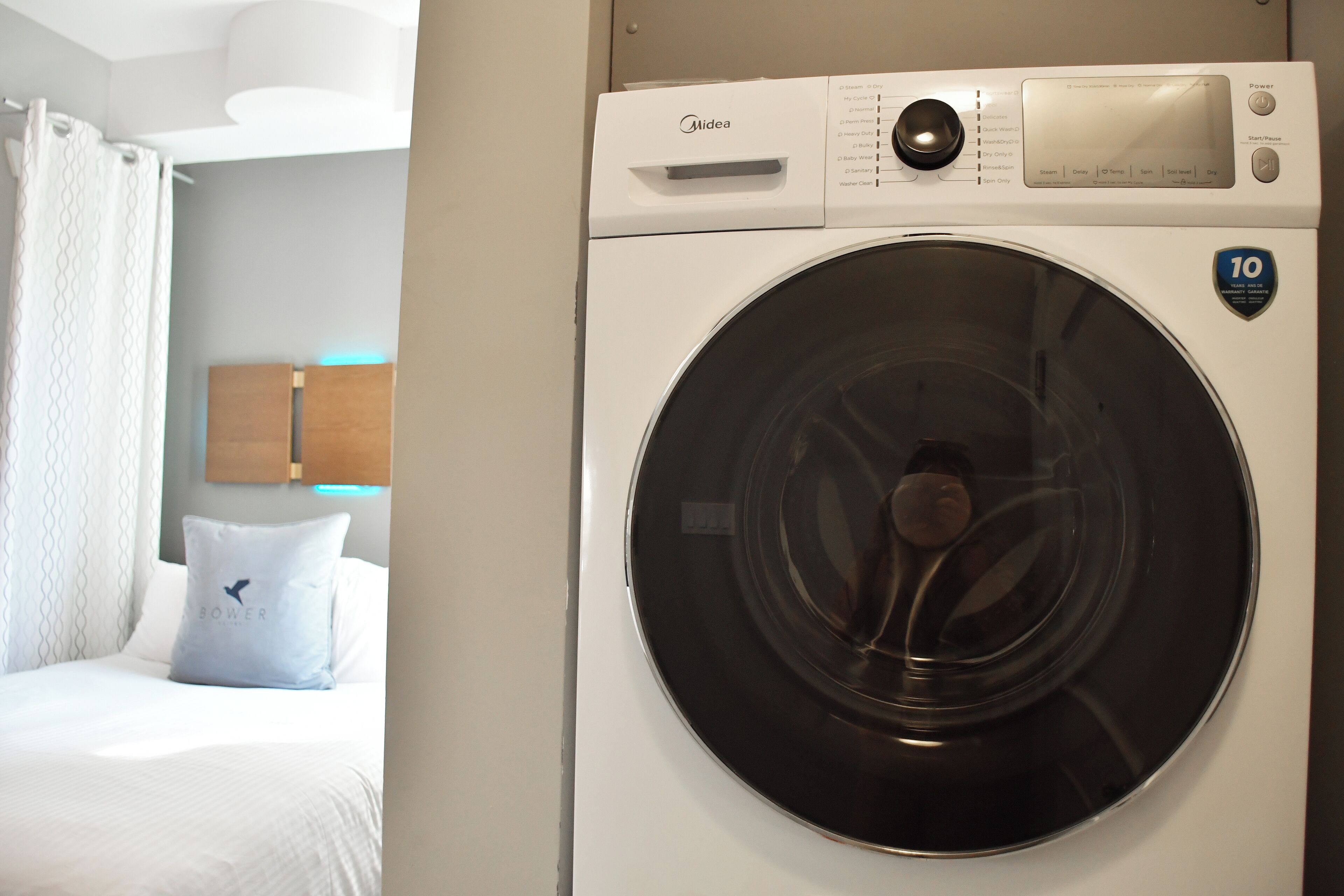 Una habitación de hotel con una lavadora Midea de carga frontal, una cama con ropa blanca y una almohada con la palabra "BOWER".