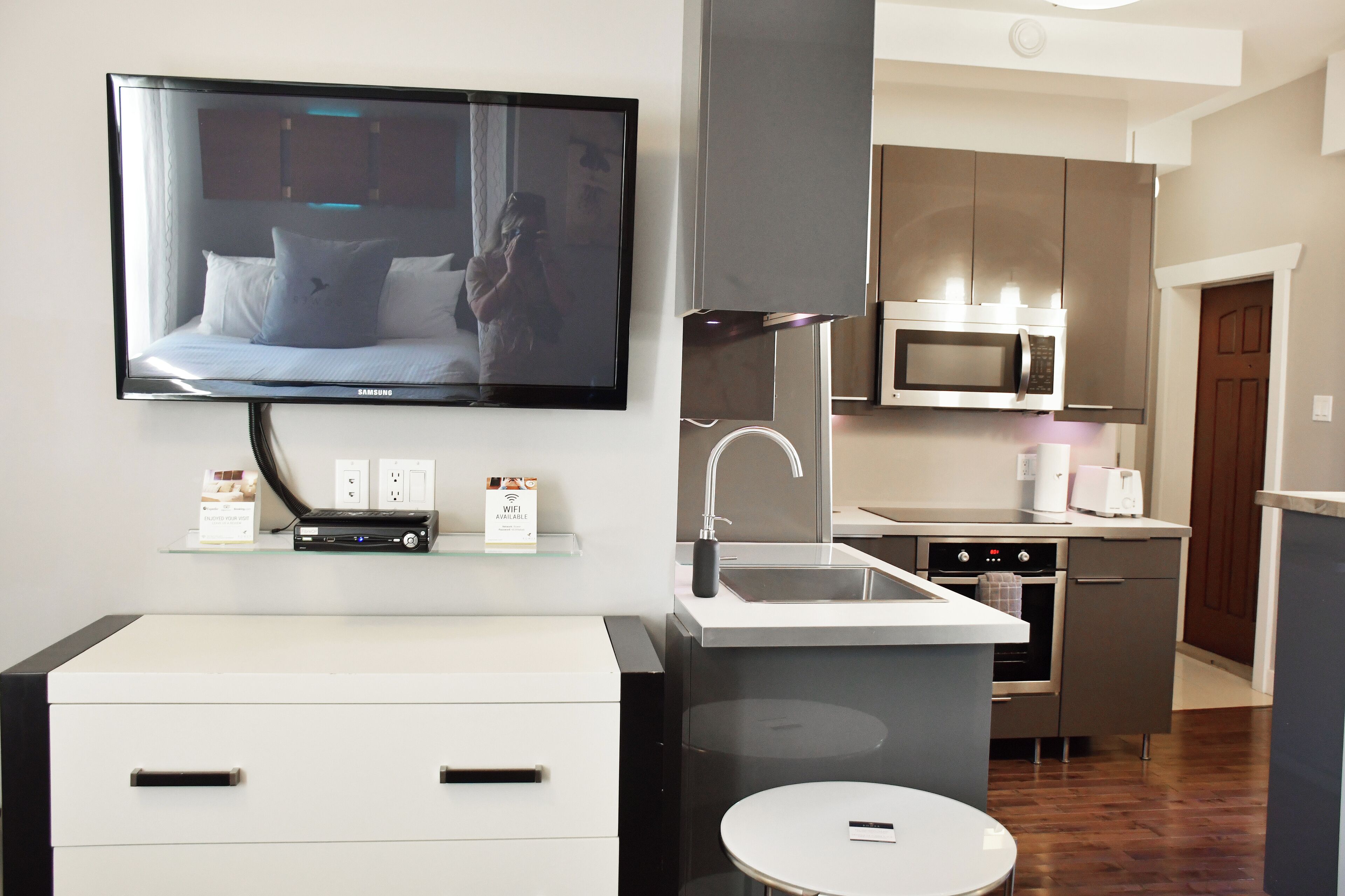 Habitación de hotel moderna con televisor de pantalla plana montado en la pared, una pequeña cocina americana con microondas, fregadero y horno, y un aparador blanco con cajones.