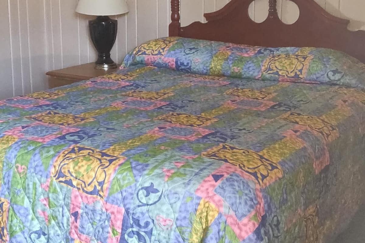 A bed with a colorful quilt, a wooden headboard, a bedside lamp, and a striped wall.
